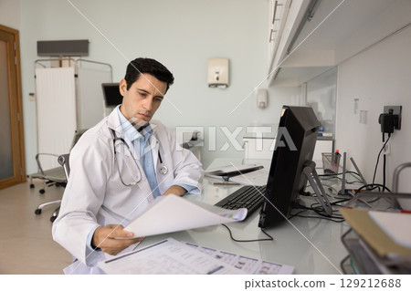 Serious young Latin doctor checking paper reports at workplace Serious young Latin doctor checking paper reports at workplace 129212688