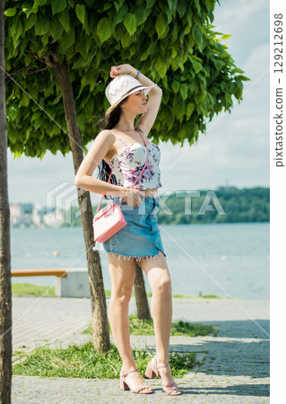 A slender girl in light clothing and a hat carries a pink bag on her shoulder while standing by the lake A slender girl in light clothing and a hat carries a pink bag on her shoulder while standing by the lake 129212698