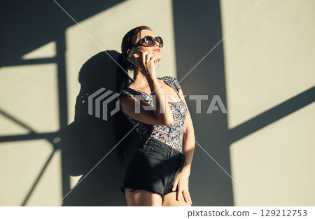 A girl in shorts and a T-shirt stands next to the light wall, talking on her cell phone. 129212753