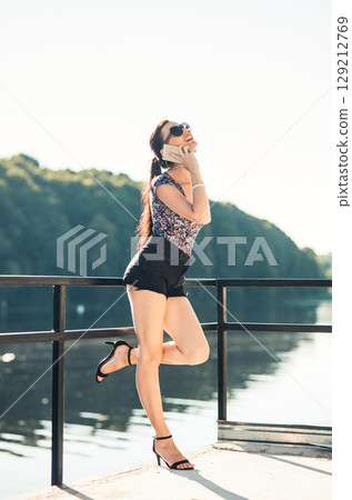 A young girl wearing short shorts and a T-shirt is talking on the phone by the lake 129212769