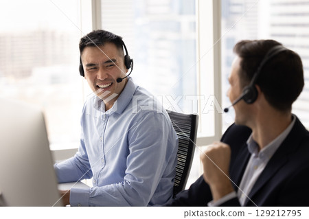 Two call center employees wear headsets laughing seated in workplace Two call center employees wear headsets laughing seated in workplace 129212795