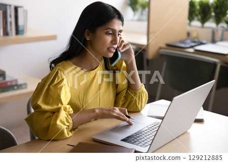 Serious Indian woman speak on phone while working on laptop Serious Indian woman speak on phone while working on laptop 129212885