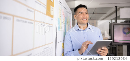 Man holding tablet stands next to whiteboard covered with notes 129212984
