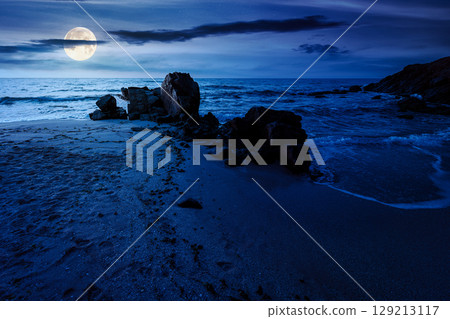 landscape by the sea at night in summer. beautiful beach with rocks washed by calm waves in full moon light. view to horizon under dark sky with clouds. scenic travel destination for leisure and relax 129213117