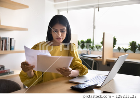 Focused woman reviewing documents sitting at table in coworking office 129213122