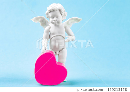 Cherub baby and pink heart on blue background. White cupid standing figurine, Valentines day greeting card Cherub baby and pink heart on blue background. White cupid standing figurine, Valentines day greeting card 129213158