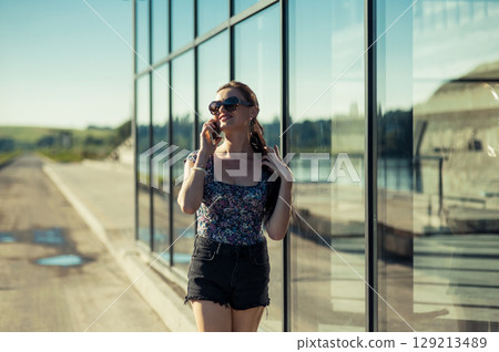 The young lady in sunglasses is wearing shorts and a T-shirt, holding the phone to her ear The young lady in sunglasses is wearing shorts and a T-shirt, holding the phone to her ear 129213489