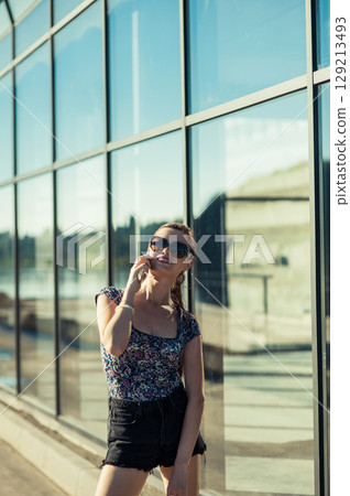 A girl wearing glasses and a ponytail is calmly talking on the phone while standing in the sun 129213493