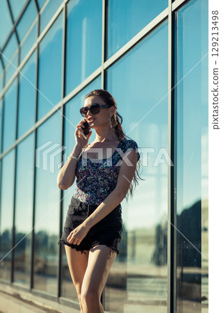 A young woman with a neat ponytail is standing near a glass building, holding the phone to her ear 129213498