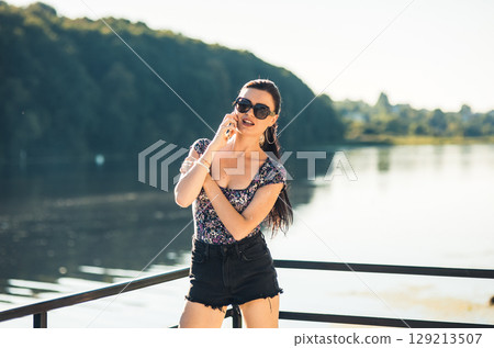 The girl with a long braided ponytail is cheerfully talking on the phone by the lake 129213507