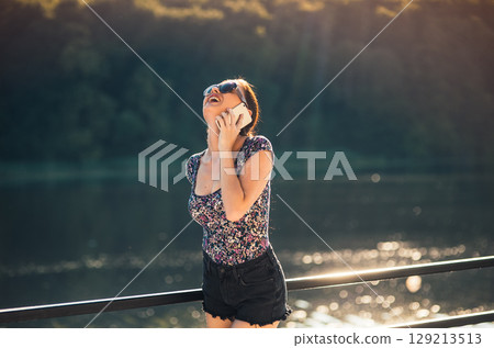 A young girl with a phone in her hand is talking and laughing as she stands by the calm water 129213513