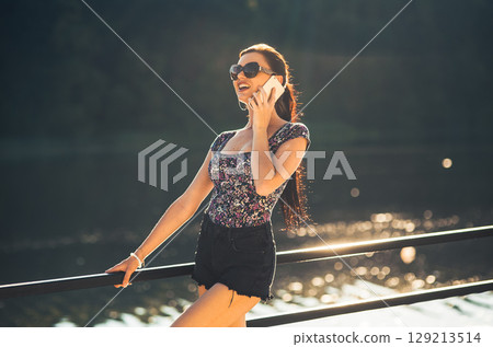 A young girl with a phone in her hand is talking and laughing as she stands by the calm water 129213514