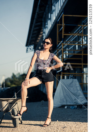 A pretty girl with her hair tied in a braid pauses next to wheelbarrows on the construction site A pretty girl with her hair tied in a braid pauses next to wheelbarrows on the construction site 129213539