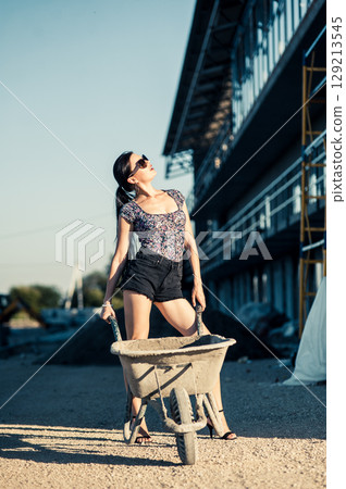 A young woman in a stylish summer outfit stands between bricks and construction wheelbarrows A young woman in a stylish summer outfit stands between bricks and construction wheelbarrows 129213545