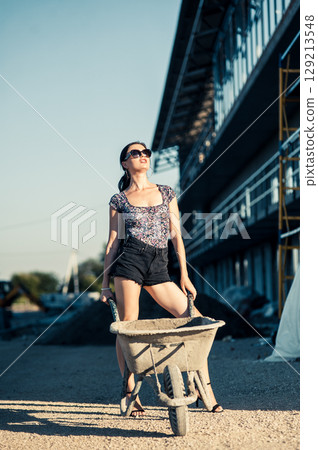 A girl in sunglasses with a braid stands near building materials, leaning slightly on a wheelbarrow 129213548