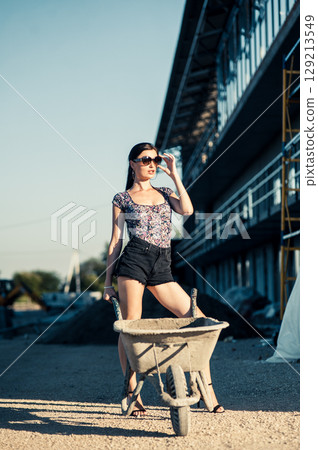 A girl in sunglasses with a braid stands near building materials, leaning slightly on a wheelbarrow A girl in sunglasses with a braid stands near building materials, leaning slightly on a wheelbarrow 129213549