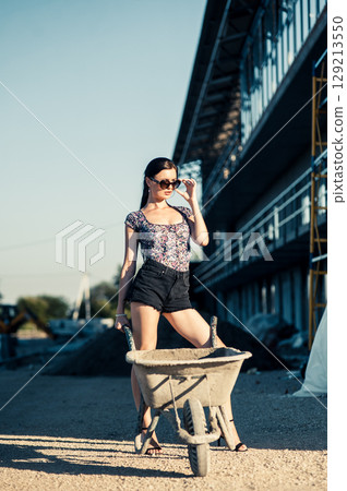 A girl in sunglasses with a braid stands near building materials, leaning slightly on a wheelbarrow 129213550