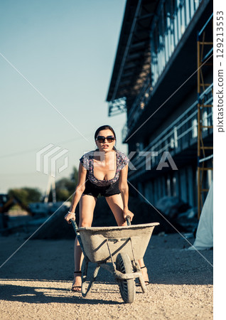 A young girl confidently poses by an old wheelbarrow in front of an unfinished building 129213553