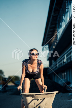 A pretty girl in summer clothes stands by scaffolding, resting her hand on the handle of a wheelbarrow 129213555