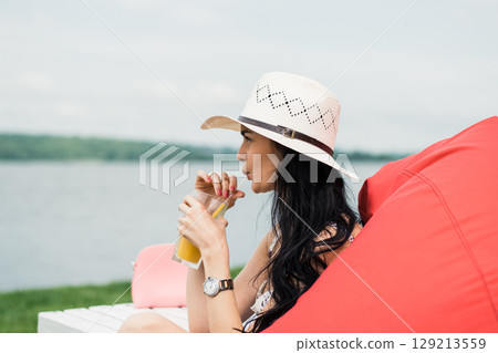 A young girl is sitting by the lake in a bright red beanbag chair, holding a glass of juice A young girl is sitting by the lake in a bright red beanbag chair, holding a glass of juice 129213559