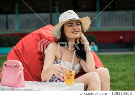 A young girl with a hat is sitting comfortably in a red beanbag chair on the grass 129213560