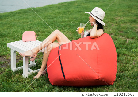 The girl in light summer clothing is nestled in a soft red chair by the lake 129213563