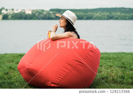A slender girl in a hat and skirt is sitting in a red chair on the grass by the water A slender girl in a hat and skirt is sitting in a red chair on the grass by the water 129213566