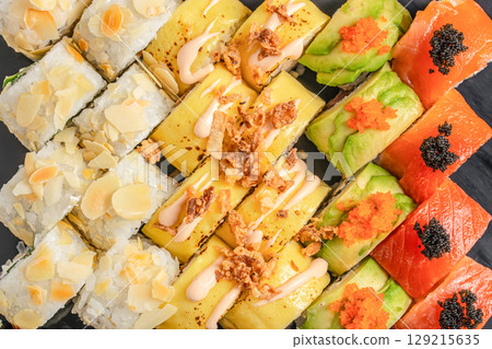 An assorted platter of vibrant sushi rolls topped with almonds, mango, avocado, and salmon with colorful roe, beautifully arranged on a dark slate background 129215635