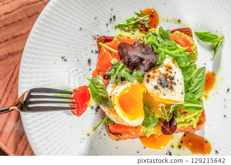 A vibrant avocado toast topped with smoked salmon, greens, and a perfectly runny poached egg, served on a white plate with a fork A vibrant avocado toast topped with smoked salmon, greens, and a perfectly runny poached egg, served on a white plate with a fork 129215642