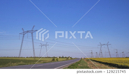 High voltage power transmission towers and power lines in a field of sunflowers under a blue sky. Power industry. Beautiful landscape. High voltage power transmission towers and power lines in a field of sunflowers under a blue sky. Power industry. Beautiful landscape. 129215745