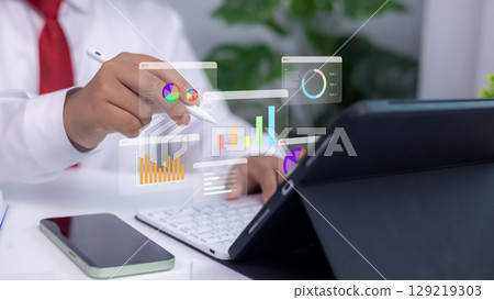 Analytics dashboards and floating holographic charts displayed as a person uses a stylus on a tablet. Concept of modern business analysis, financial planning, and data-driven technology. 129219303