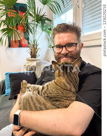 Happy man holding his cat on a couch at home. Companionship, relaxation, and emotional comfort through the bond between human and pet. Happy man holding his cat on a couch at home. Companionship, relaxation, and emotional comfort through the bond between human and pet. 129220258