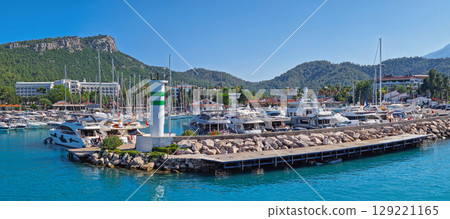 KEMER, TURKEY - JULY 3, 2025 Marina panoramic view with Calis Hill in the background. Rocky pier filled with numerous boats and yachts on the clear turquoise water under a bright blue Turkish sky 129221165
