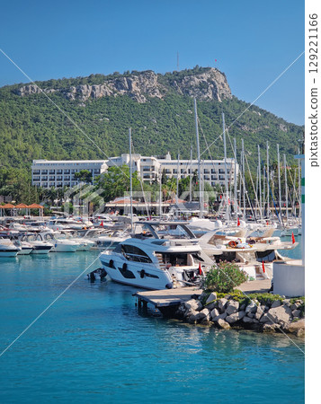 Kemer marina filled with numerous boats and yachts with Calis Hill in the background. A lighthouse stands prominently on the rocky pier, overlooking the clear blue water under a bright blue sky Kemer marina filled with numerous boats and yachts with Calis Hill in the background. A lighthouse stands prominently on the rocky pier, overlooking the clear blue water under a bright blue sky 129221166