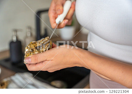 Close up view of female hands struggling to open a fresh oyster at home with a special oyster knife. Risks and challenges of handling shellfish without professional training 129221304