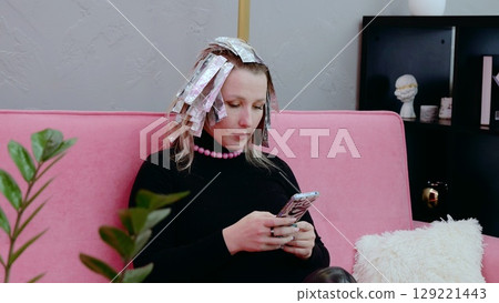 Young woman with foil in her hair uses a smartphone while waiting for her hair dye to set in at a beauty salon, relaxing on a pink sofa Young woman with foil in her hair uses a smartphone while waiting for her hair dye to set in at a beauty salon, relaxing on a pink sofa 129221443