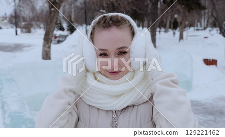 Winter scene featuring smiling young woman wearing white earmuffs and warm scarf, standing amid snow covered park landscape, holding protective ear coverings 129221462