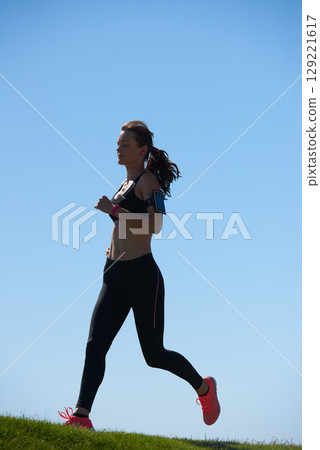 Woman runner, grass and sky for fitness, health and wellness in summer, sunshine or outdoor. Athlete girl, running and workout with tech, phone or exercise for speed, self care or healthy strong body 129221617