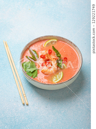 Traditional Thai soup tom yum soup with shrimp, lime, and herbs in bowl with chopsticks on blue background 129221749