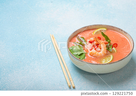 Traditional Thai soup tom yum soup with shrimp, lime, and herbs in bowl with chopsticks on blue background, space for text 129221750