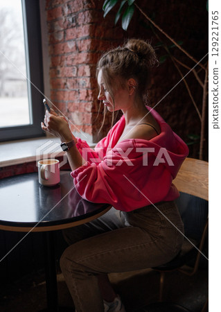 A stylish woman enjoying her coffee while scrolling on her phone in a warm cafe setting. A stylish woman enjoying her coffee while scrolling on her phone in a warm cafe setting. 129221765