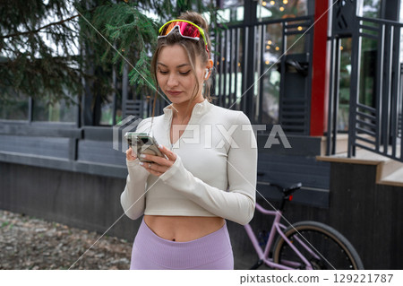 A woman in stylish activewear checks her phone, showcasing the vibrant outdoor fitness culture by her bicycle. 129221787