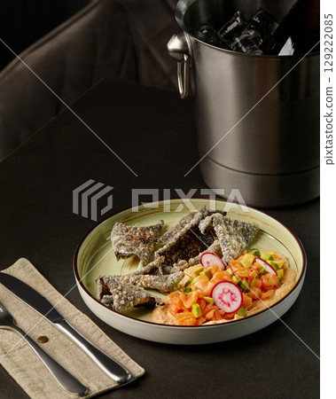 Salmon tartare with avocado, radish, and crispy nori chips on a table with cutlery and ice bucket 129222085