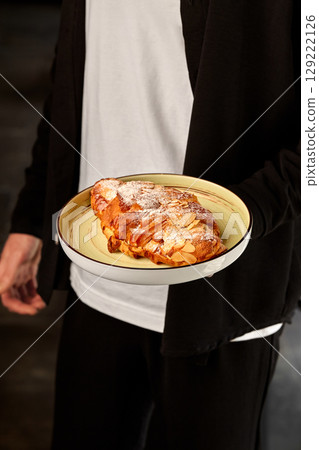 Baker holding freshly baked almond croissant on plate 129222126