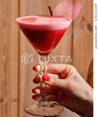 Woman hand holding elegant glass with red layered cocktail decorated with pink leaf 129222204