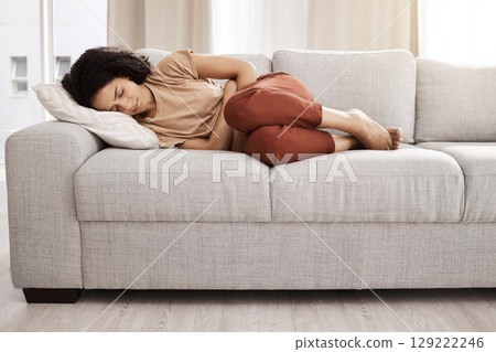 Black woman, stomach pain and digestion with constipation, problem and endometriosis on the sofa in home. Young female, sick and abdomen cramp and diarrhea with ibs, menstrual cycle and bowel ache 129222246