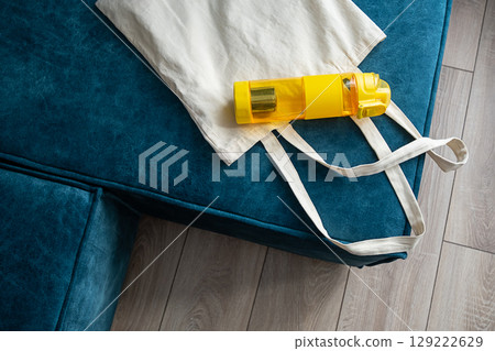 Reusable water bottle and fabric shopper bag on a blue sofa. 129222629
