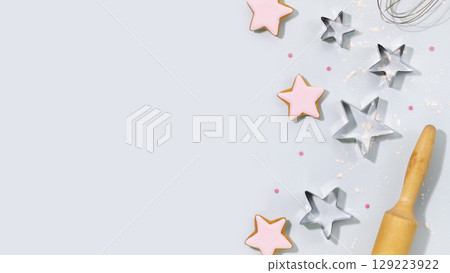 Cooking holiday cookies. Bakery kid background. Ginger cookies in the form of stars, covered with glaze, cookie cutters, a dough gurney and a whisk on a blue background. Top view, copy space 129223922