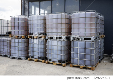 A stack of large, rectangular, metal-framed containers filled with various colored liquids are positioned on wooden pallets in an outdoor industrial setting 129224807