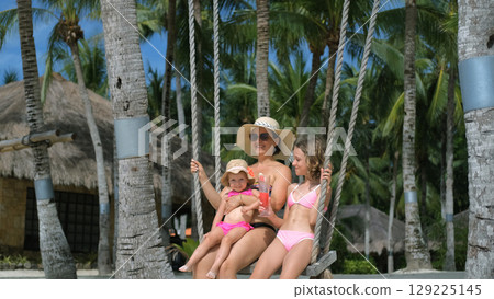 Young mother swings with daughters on tropical beach while older girl sips watermelon shake 129225145